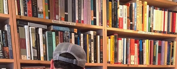 The top of a head, wearing a baseball cap, set against a backdrop of rows of books in a bookshop